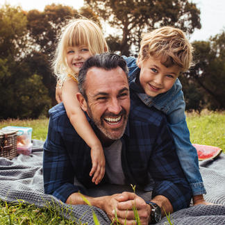 Father laughing with two children on picnic blanket outdoors, with picnic basket and fruit visible in background.