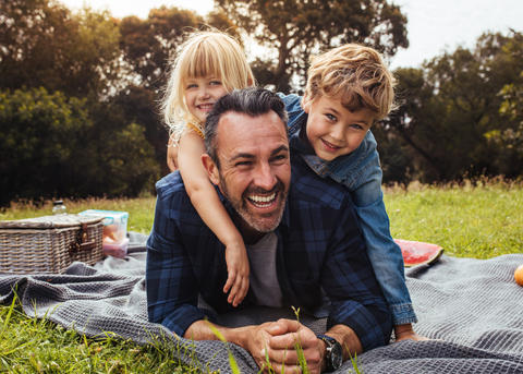 Father laughing with two children on picnic blanket outdoors, with picnic basket and fruit visible in background.