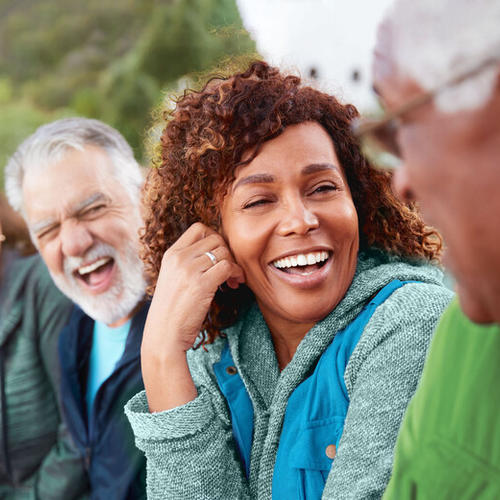 Four older adults sitting outdoors together, laughing and enjoying a relaxed moment in a natural setting while wearing casual clothes.